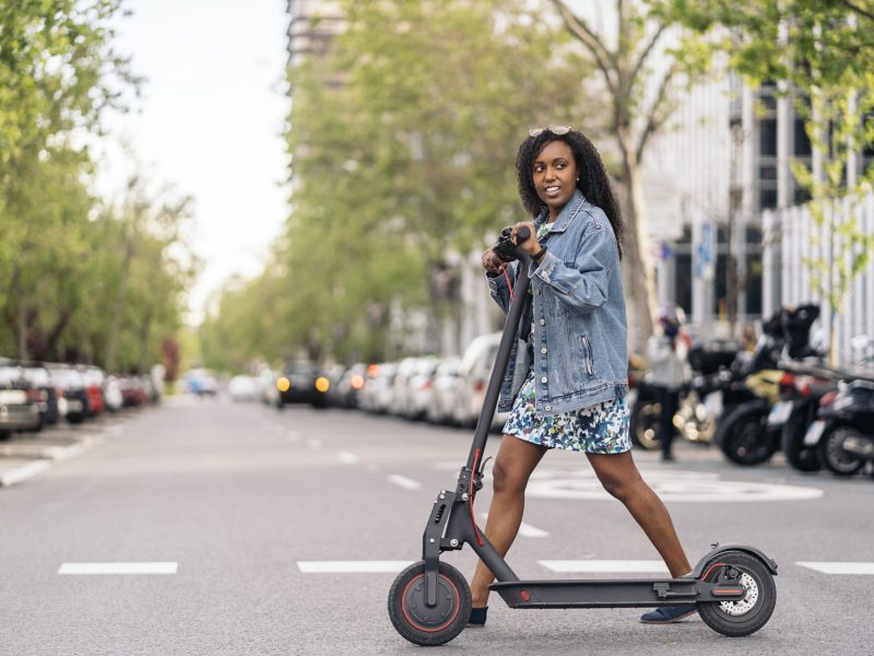 Young black girl wearing colorful dress crossing the street with her electric scooter during sunny day.